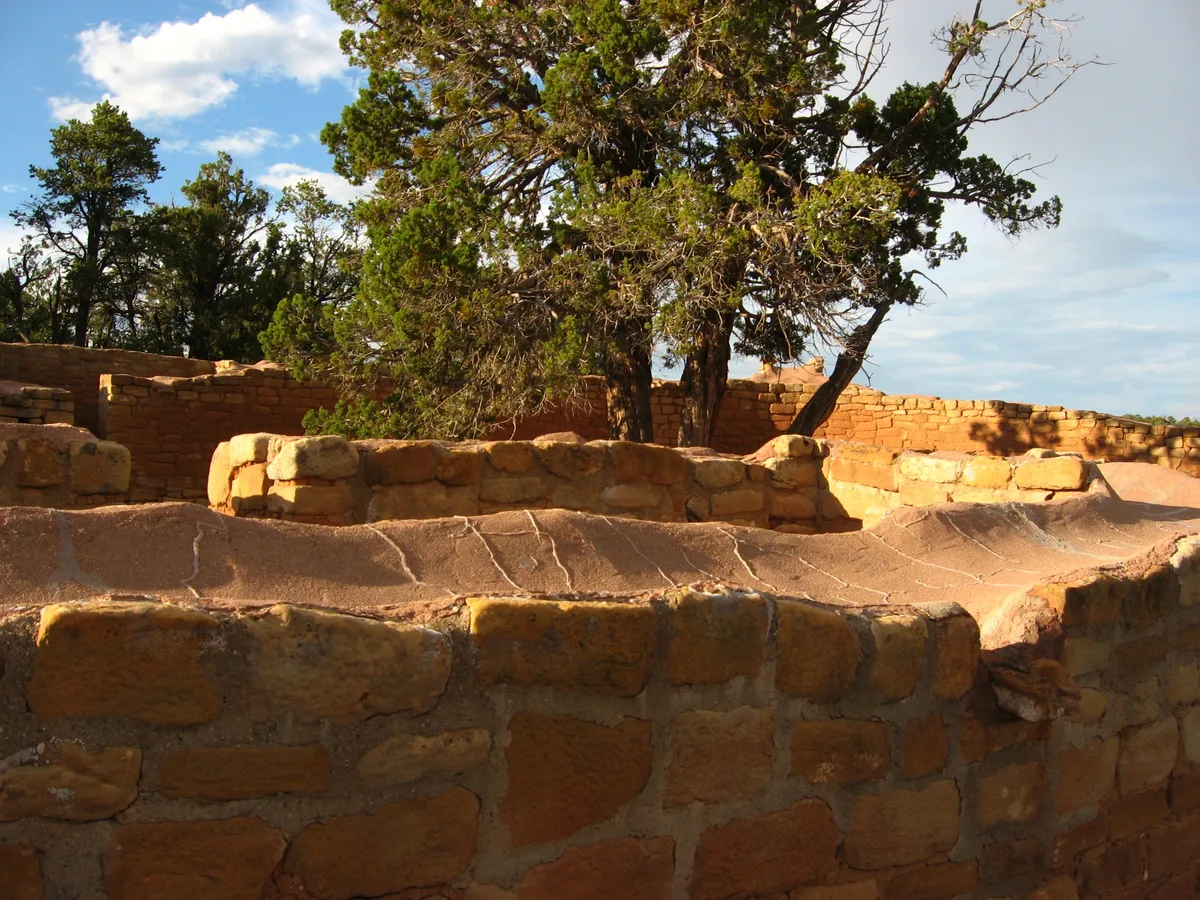 Sun Temple’s curved sandstone walls fill the foreground, their sunlit masonry glowing warm gold and red beneath a low afternoon sky. Pinyon trees rise from the unfinished ceremonial enclosure, with the D-shaped stonework and layered wall courses clearly visible.
