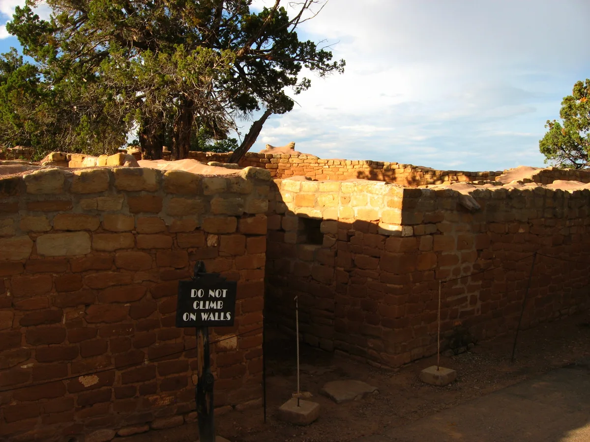 Sun Temple’s reddish stone walls fill the foreground, with a dark doorway opening into the unfinished masonry and a “DO NOT CLIMB ON WALLS” sign set in front. Low afternoon light warms the blocks while juniper trees and a bright blue sky rise above the mesa-top ruin.