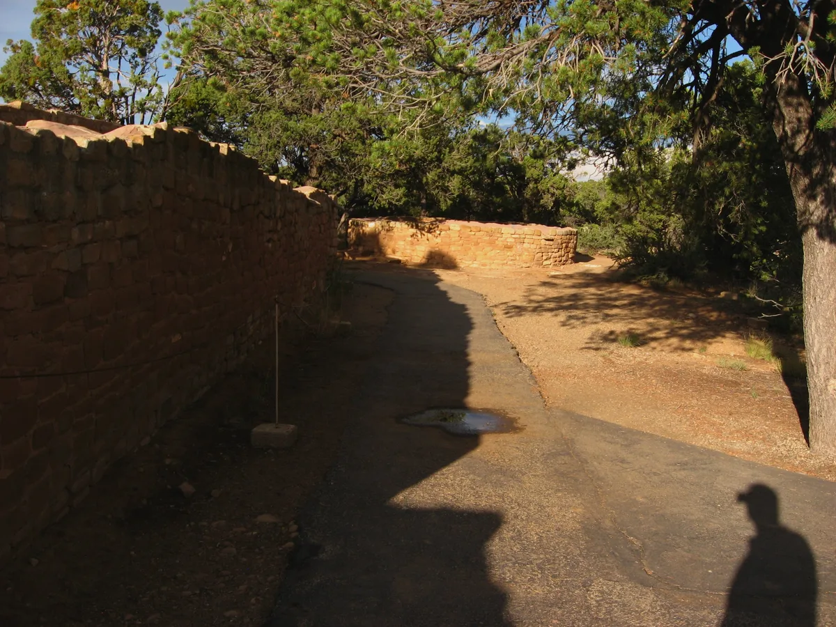 Sunlit sandstone masonry walls curve along a paved path at Sun Temple, with deep shadows from nearby pines falling across the ground. The unfinished ceremonial structure sits on dry mesa-top terrain, framed by trees and warm afternoon light.