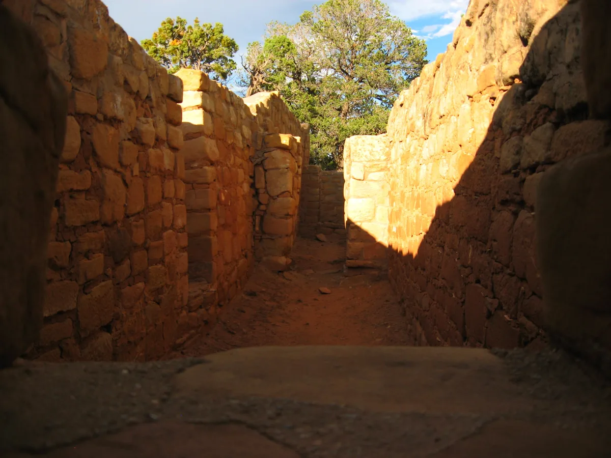 Sunlit sandstone masonry walls form a narrow passage through the unfinished Sun Temple, with rough stone blocks and earthen floor leading toward a small opening. Green trees and blue sky rise above the structure, while warm afternoon light casts deep shadows across the corridor.
