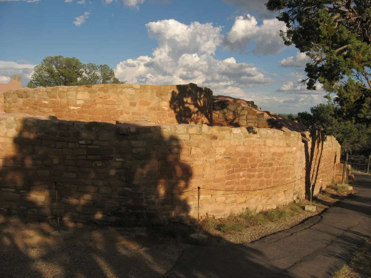 Sun Temple’s curved sandstone masonry fills the frame, its low D-shaped wall glowing in warm afternoon light and cast with tree shadows. A paved path and rope barrier run along the right edge, with pine branches and billowing clouds above the mesa-top site.