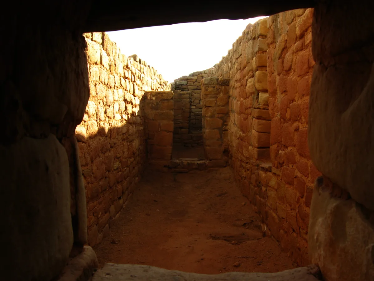 Sunlight spills into a narrow stone corridor inside Sun Temple, highlighting the rough tan masonry walls and the packed dirt floor. The dark foreground frames the passage, which ends at a small opening and a partially built stone wall.