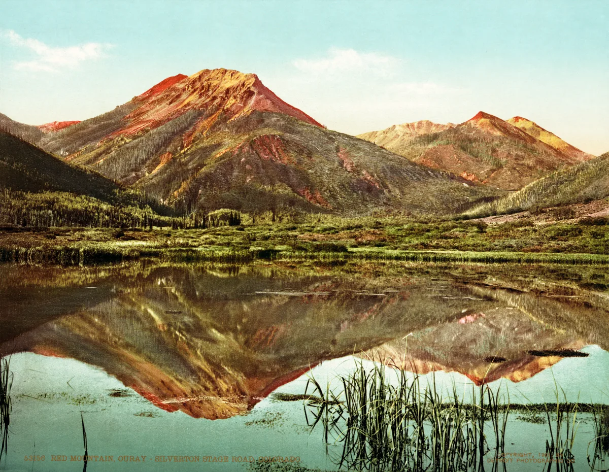 Red Mountain and nearby peaks glow in streaks of red, orange, and gold above a still alpine pond that mirrors the mountains almost perfectly. Tufts of reeds break the water in the foreground, and the wide valley under a pale blue sky emphasizes the stark, colorful terrain.