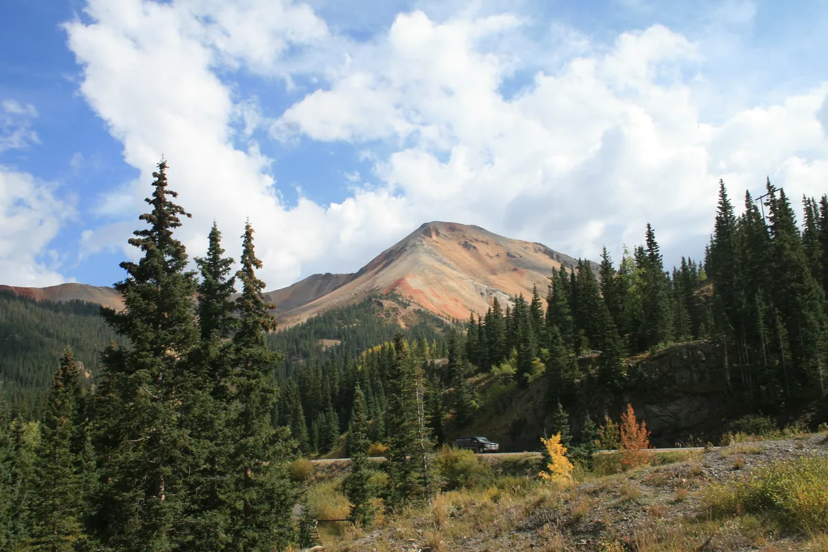 A wide mountain overlook shows a red-and-tan alpine peak rising above dark evergreen forests, with the Million Dollar Highway curving through the valley below. Puffy white clouds fill the bright afternoon sky, and a few yellow-orange shrubs add color in the foreground.