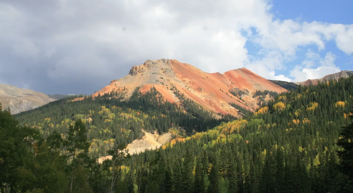 Sunlit red and orange peaks rise above a dense forest of dark evergreens with patches of yellow autumn color, under a partly cloudy blue sky. The scene captures the steep, barren slopes and vivid mineral-stained rock of Red Mountain Pass.