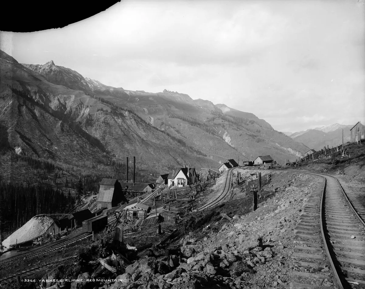 A steep mountain mining site with a narrow railroad track curving along the hillside past small wooden buildings, ore bins, and scattered timbers. Jagged alpine peaks rise in the background under a bright, hazy sky, giving the black-and-white scene a stark, high-elevation feel.
