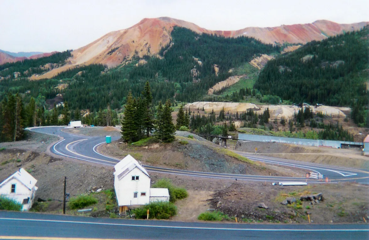 Winding lanes of the Million Dollar Highway curve through a barren alpine pass, with small white mining buildings in the foreground and dark evergreens climbing the slopes. Behind them, the Red Mountain peaks rise in vivid bands of red, orange, and tan under soft afternoon light.
