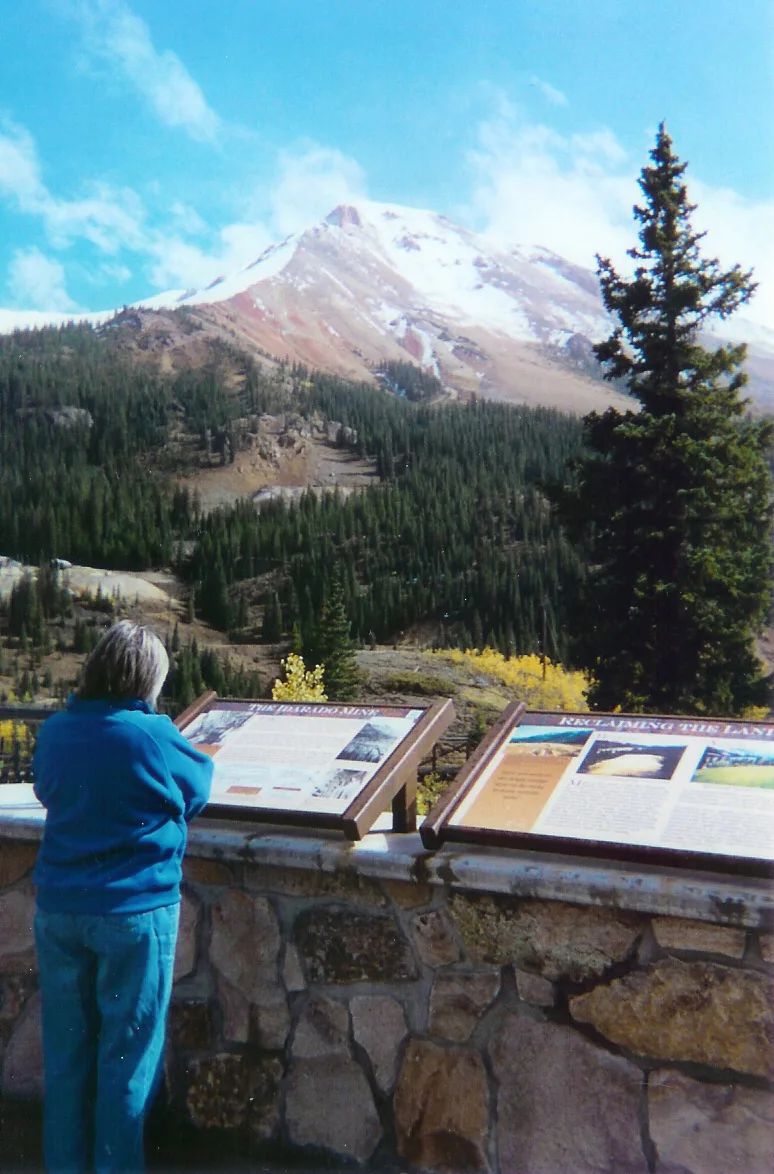 A visitor in a bright blue jacket reads interpretive signs at a stone overlook while a snow-dusted mountain rises behind a dark evergreen forest. The afternoon light picks out patches of red and tan rock on the slope under a vivid blue sky.