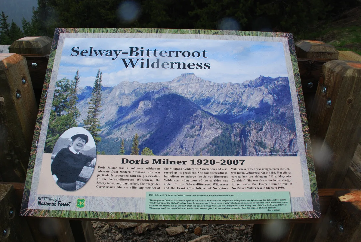 A dedication sign for Doris Milner stands on a wooden post, showing a large mountain photograph labeled “Selway-Bitterroot Wilderness” above her portrait and memorial text. Evergreen trees and a dark forested slope frame the sign in the background.