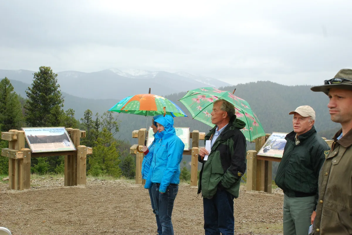 Several people stand on a misty mountain overlook in rain jackets, holding colorful umbrellas, with the Continental Divide signposts and interpretive plaques behind them. Snow-dusted ridges fade into the cloudy background beyond the pine-covered slopes.