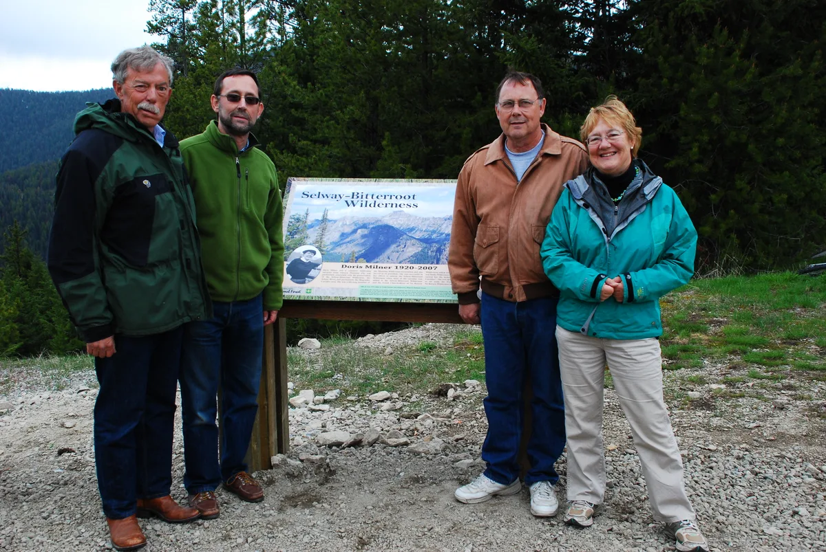 Four people stand beside a trailhead sign for the Selway-Bitterroot Wilderness, with dark evergreens and a forested mountain slope behind them. The rocky turnout and cool, overcast light give the high-elevation scene a crisp, subdued look.