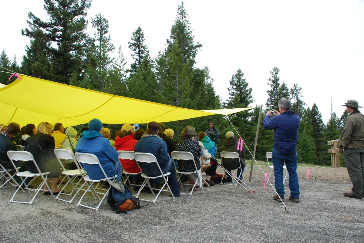 People sit on white folding chairs under a bright yellow tarp at a high mountain overlook, listening to a speaker with evergreen trees behind them. A man in blue photographs the scene while another person stands near a wooden signpost at the edge of the gravel clearing.