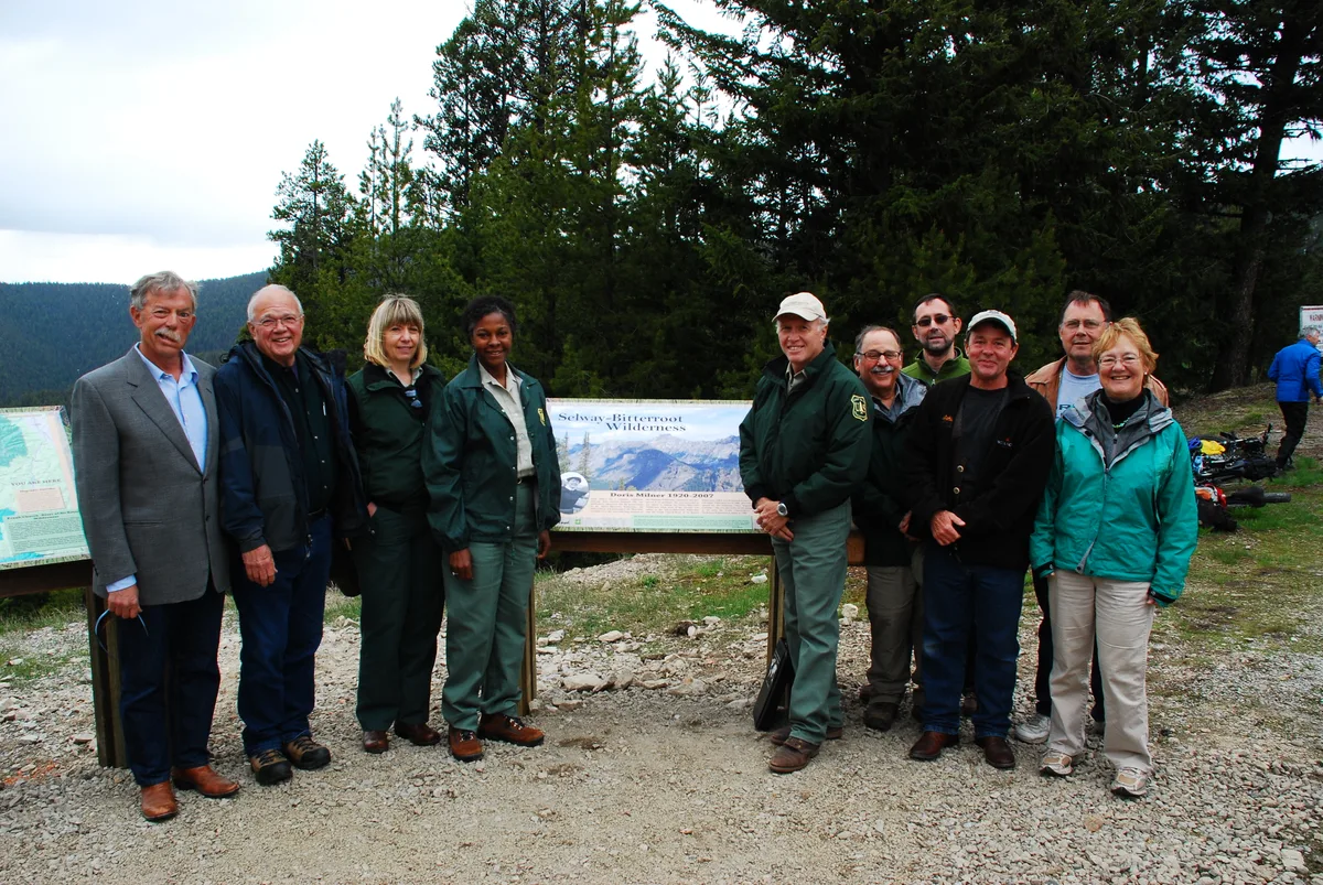 A group of nine people stands on a gravel overlook beside interpretive signs at Milner Pass, with dense evergreen forest and a mountain ridge rising behind them. One sign reads “Selway-Bitterroot Wilderness,” and the overcast light gives the scene a cool, muted tone.