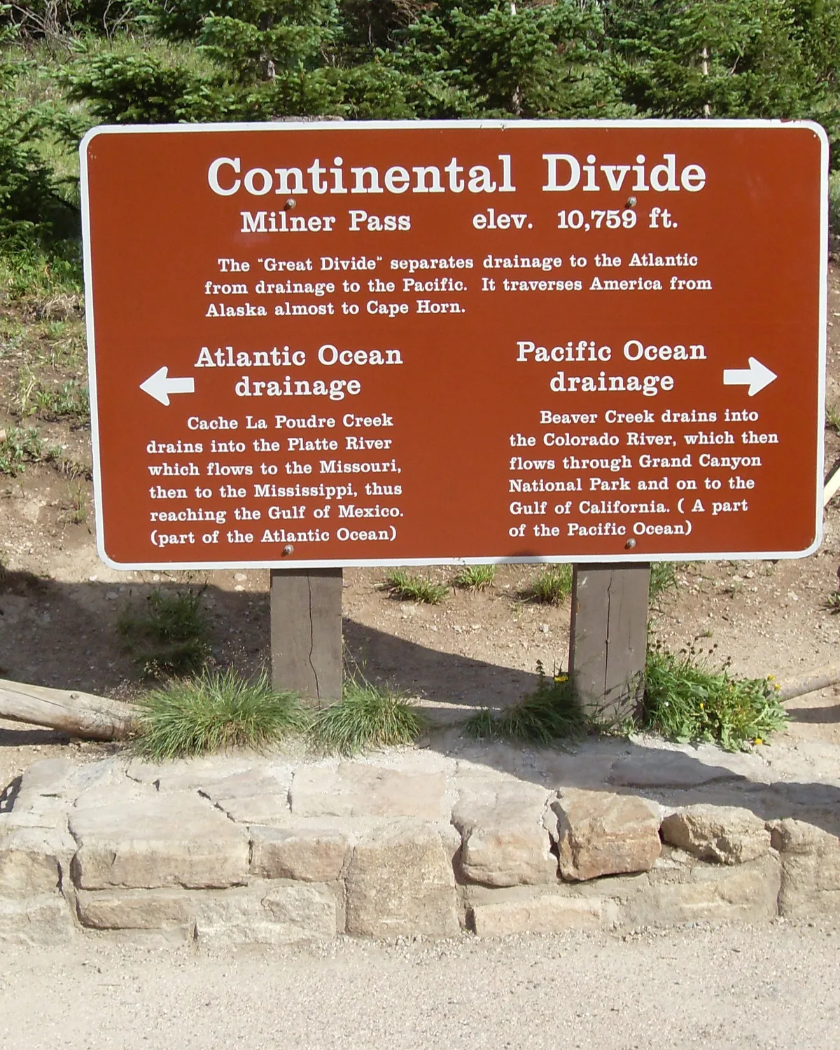 A brown Continental Divide sign at Milner Pass stands in front of evergreen trees and rocky ground, with white arrows pointing to Atlantic Ocean drainage on the left and Pacific Ocean drainage on the right. The sign marks the 10,759-foot pass and explains how water flows in opposite directions from the divide.