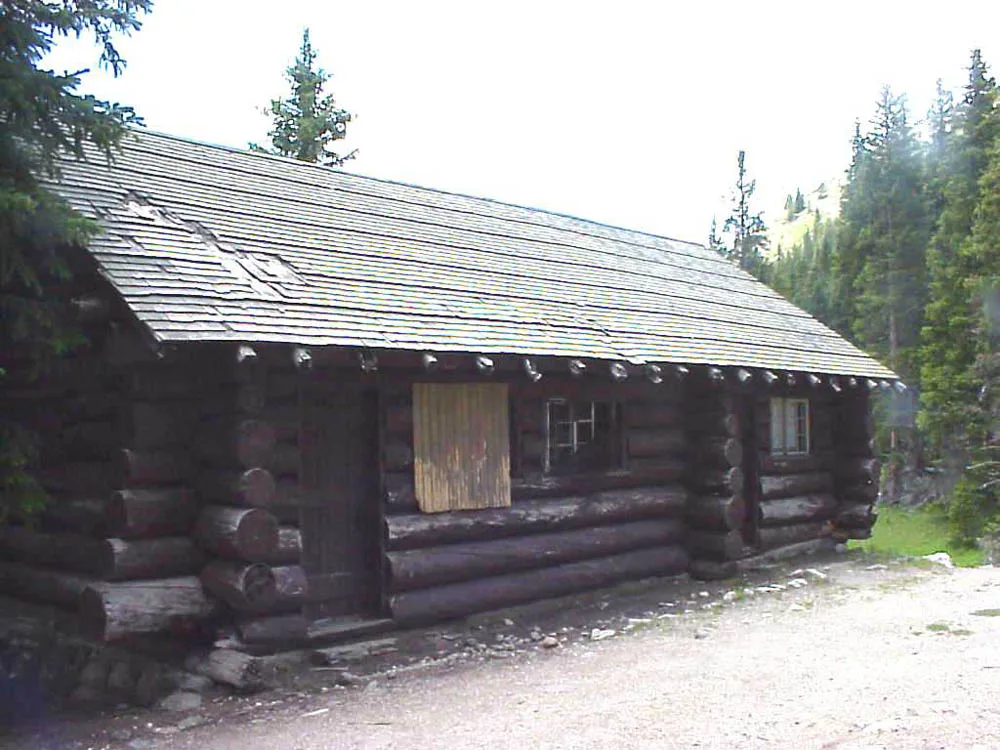 A rustic log cabin with a steep shingled roof sits beside a gravel road, partly shaded by tall evergreens. Bright sunlight washes the treetops and the pale sky, while the dark timber walls and small windows give the building a historic mountain-camp feel.