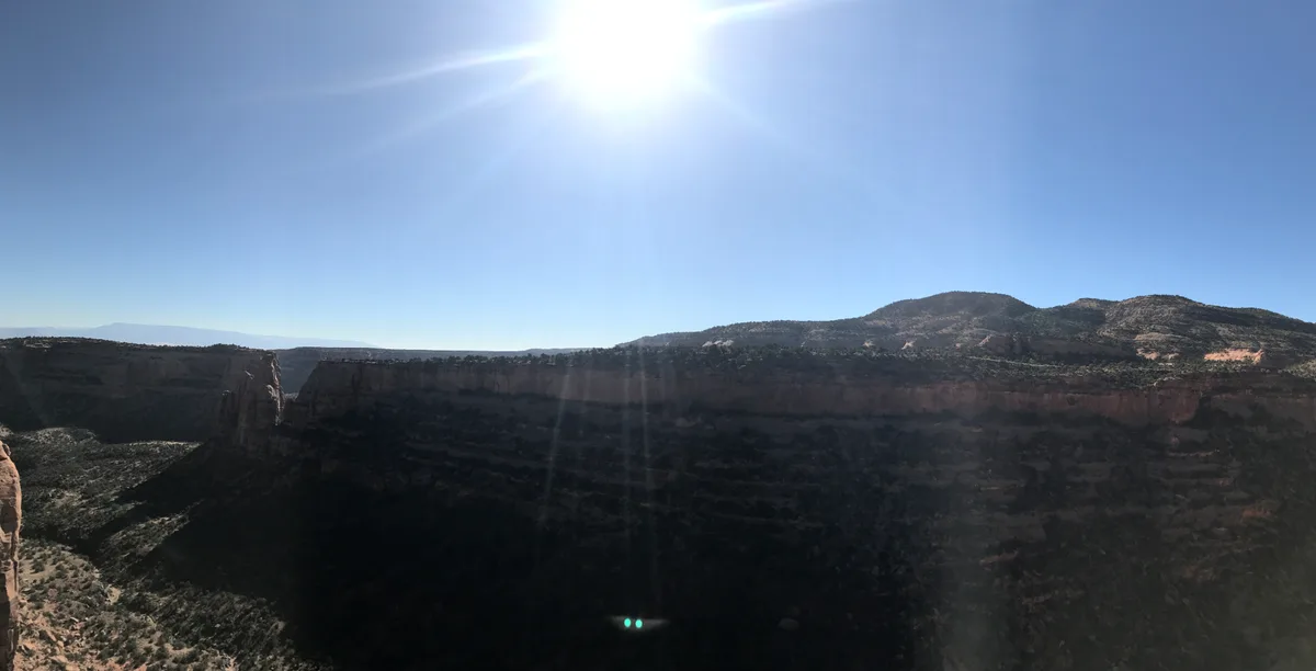 Bright morning sun hangs high over a wide desert canyon, casting the foreground into deep shadow while sunbeams and lens flare streak down across the dark sandstone cliffs. Low, scrub-covered mesas line the horizon beneath a clear blue sky.
