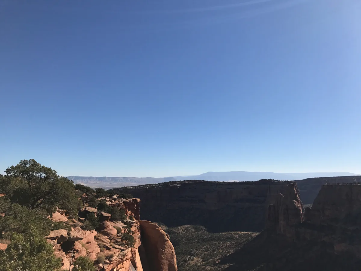 Sunlit red sandstone cliffs and a narrow balanced rock formation rise from a dry canyon, with scrubby evergreens clinging to the rocky rim. A vast blue sky fills most of the frame, emphasizing the open desert landscape and distant mesas.