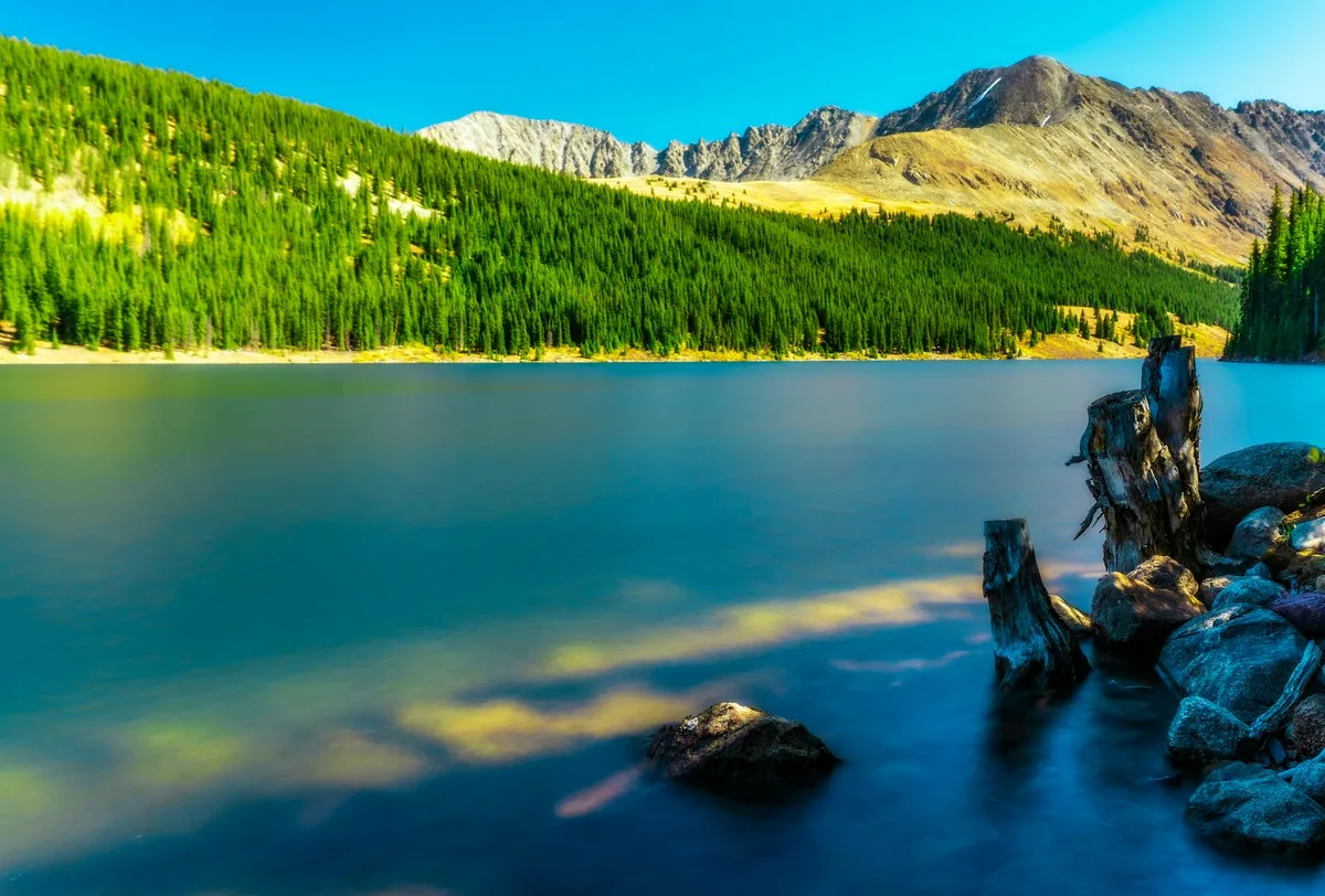 Long-exposure view of a turquoise alpine lake with smooth, misty water in the foreground and sunlit evergreen slopes rising to rugged mountain peaks under a clear blue sky. Weathered tree stumps and rocks line the right shore, adding texture to the serene mountain scene.