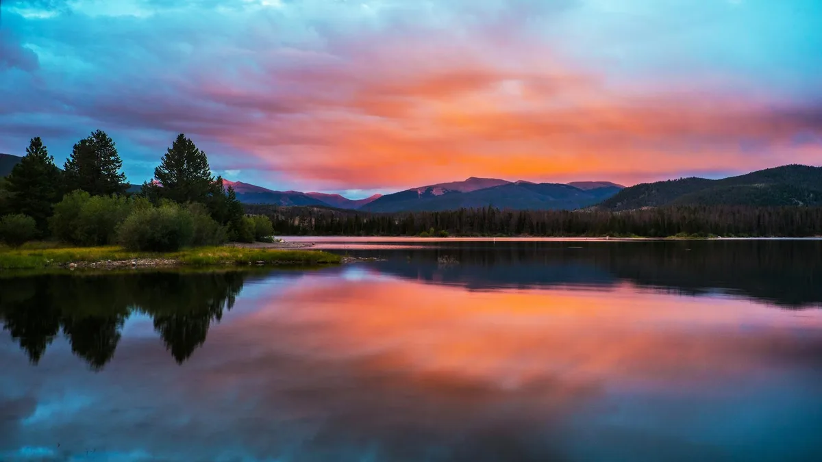 A calm alpine lake mirrors a vivid sunrise sky streaked with pink, orange, and blue, with dark pine-covered hills and mountains lining the horizon. The smooth water creates a near-symmetrical reflection, while a small wooded shoreline anchors the left side of the frame.