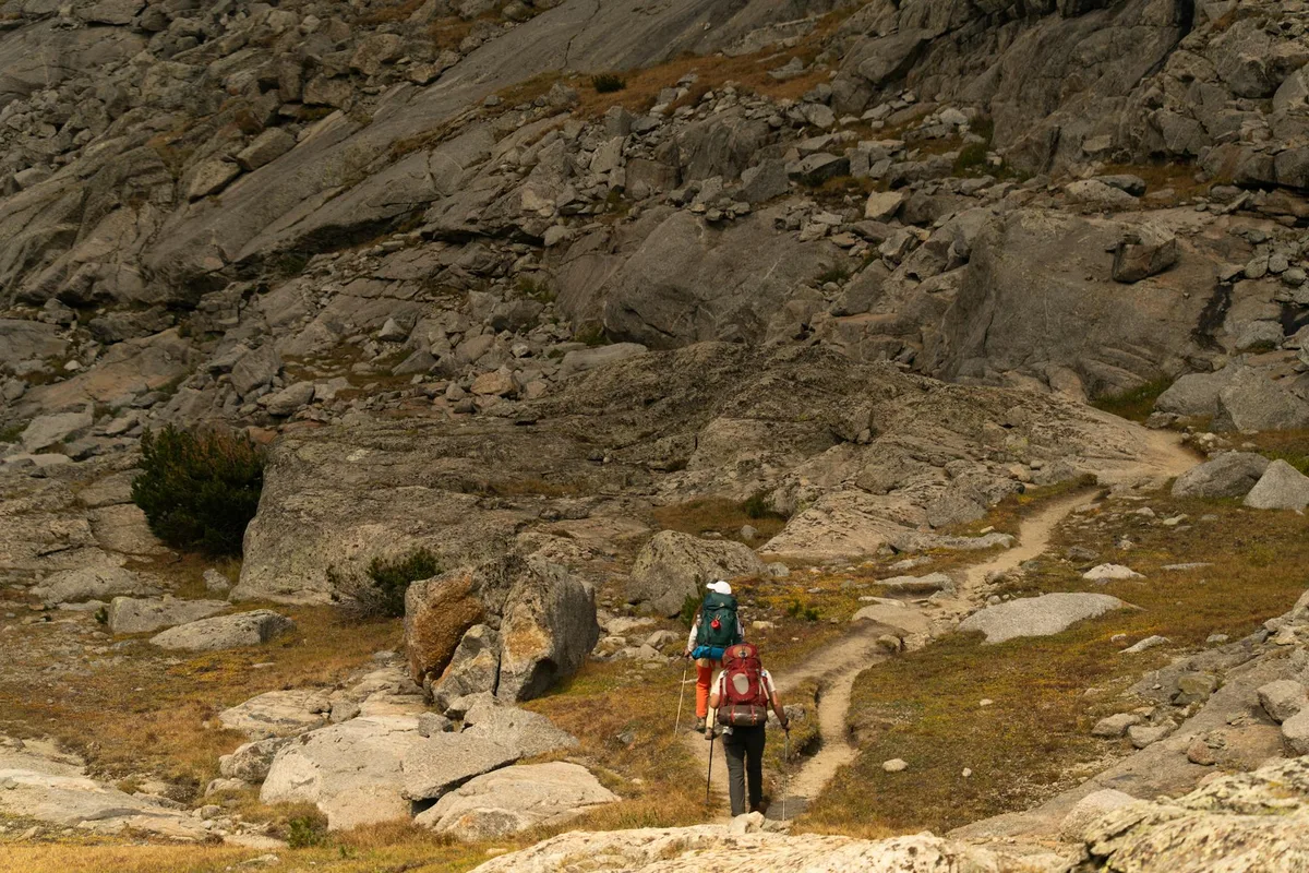 Two hikers with backpacks and trekking poles follow a narrow dirt trail across a rugged alpine slope, surrounded by boulders, granite slabs, and patches of sparse high-country grass. The morning light is soft and muted, emphasizing the rocky texture and open, remote mountain landscape.