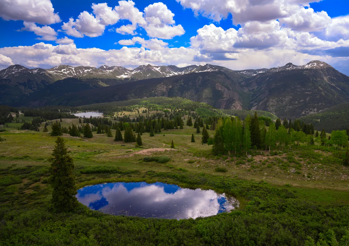 Molas Lake reflects a patch of bright blue sky and towering cumulus clouds in the foreground, with rolling alpine meadows and dark evergreen stands stretching toward the snow-streaked Grenadier Range. The wide view emphasizes the dramatic mountain skyline and the vivid green slopes in golden light.