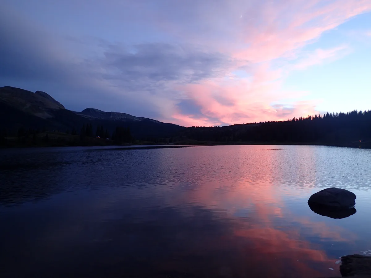 Molas Lake is calm at sunset, reflecting pink and blue clouds across the water with dark mountain ridges and a tree-lined horizon in silhouette. A few rocks break the mirrorlike foreground, adding to the quiet alpine scene.