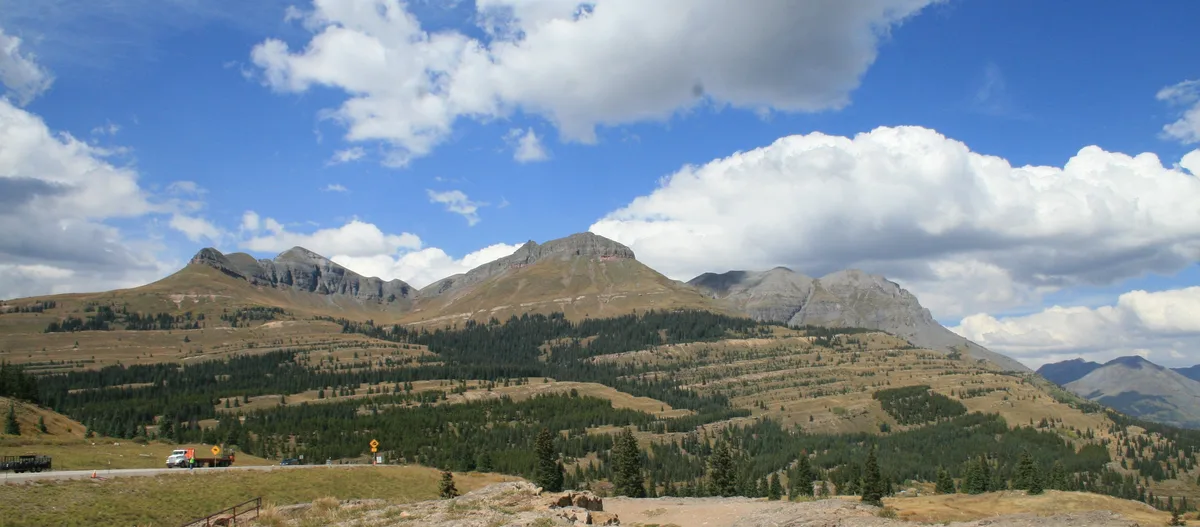 Sweeping alpine overlook with the Grenadier Range rising across a broad, forested valley, under a bright blue sky filled with large, sunlit clouds. In the foreground, a roadside turnout and low rocky slope frame the expansive mountain view.