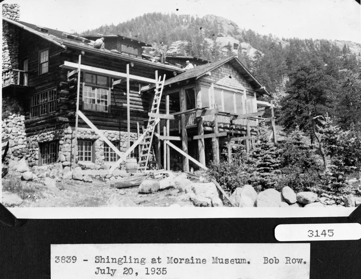 A black-and-white historic photo shows a rustic stone-and-log museum building under construction, with workers on the roof, a ladder propped against the facade, and timber scaffolding around the porch. Pine-covered mountains rise behind the lodge-like structure, with rough boulders and shrubs in the foreground.