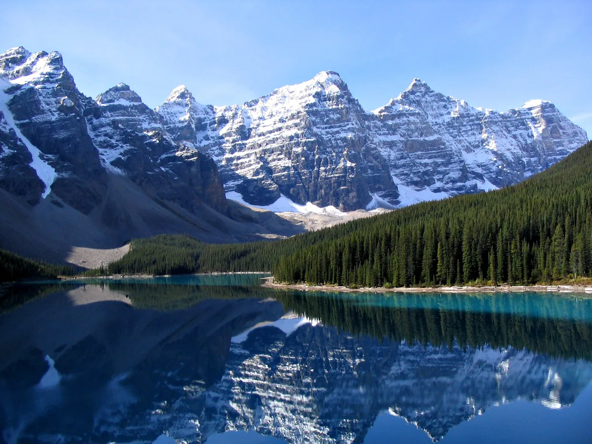 Snow-capped Rocky Mountain peaks rise above a calm alpine lake, with dense evergreen forest lining the shore. The still water creates a near-perfect reflection of the mountains and blue sky, emphasizing the clear, bright light.