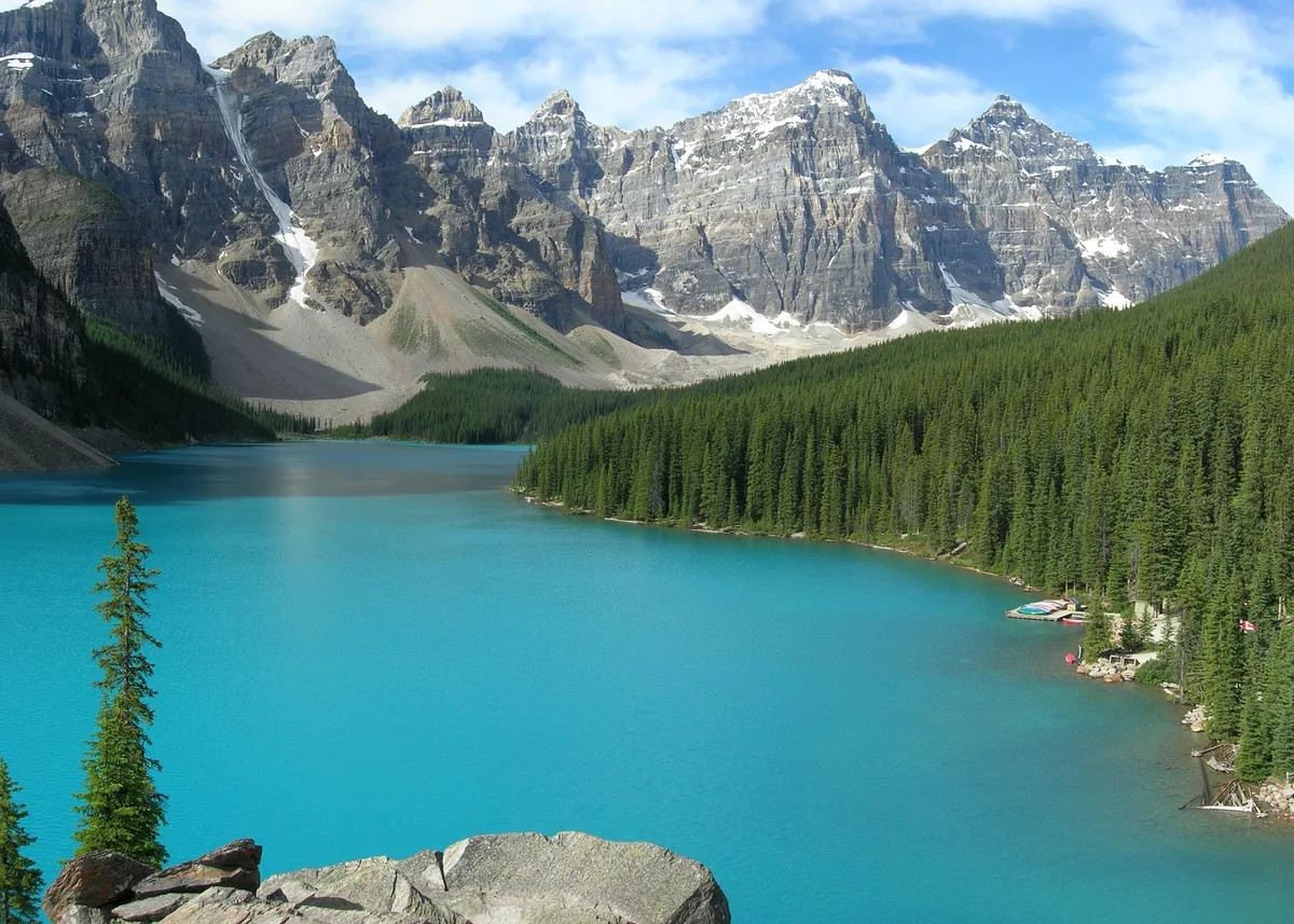 A vivid turquoise mountain lake fills the foreground, edged by a dense evergreen forest and backed by steep gray peaks streaked with lingering snow. The bright blue sky and crisp light give the scene a clear, alpine feel.