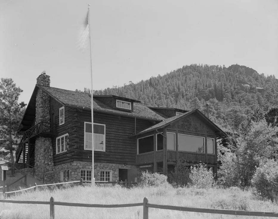 A rustic stone-and-log museum building sits in a grassy meadow with a flagpole in front, backed by a forested mountainside under a clear sky. The low, black-and-white composition highlights the cabin-like architecture and surrounding trees more than the distant landscape.