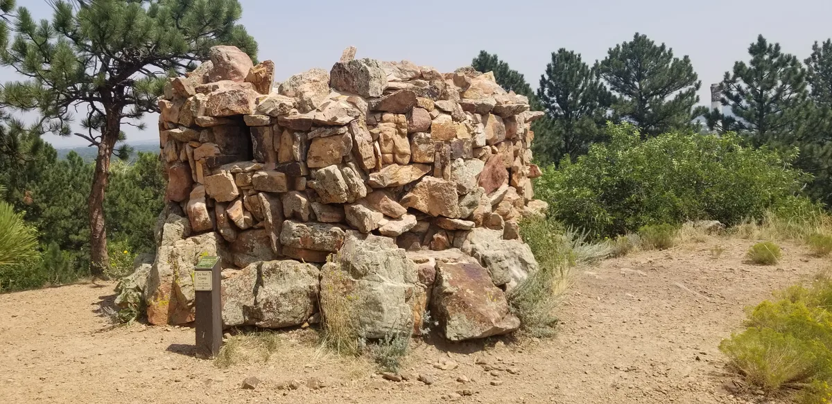 A rough stone summit shelter sits on a dusty mountaintop, framed by pine trees and scrub with hazy sky beyond. The tan and pink boulders catch soft daylight, giving the scene a warm, rugged feel.