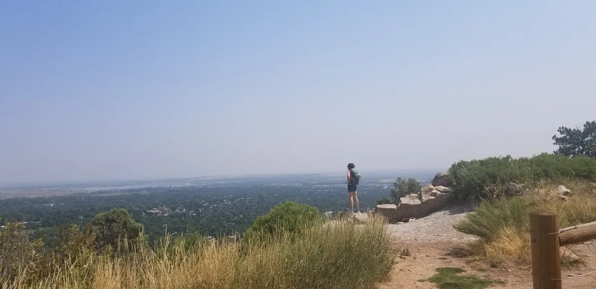A lone hiker stands on the rocky Mount Sanitas summit overlook, gazing out over hazy Boulder and the flat plains beyond. Dry grasses, scrub, and pale quartzite boulders frame the wide panoramic view under soft golden-hour light.