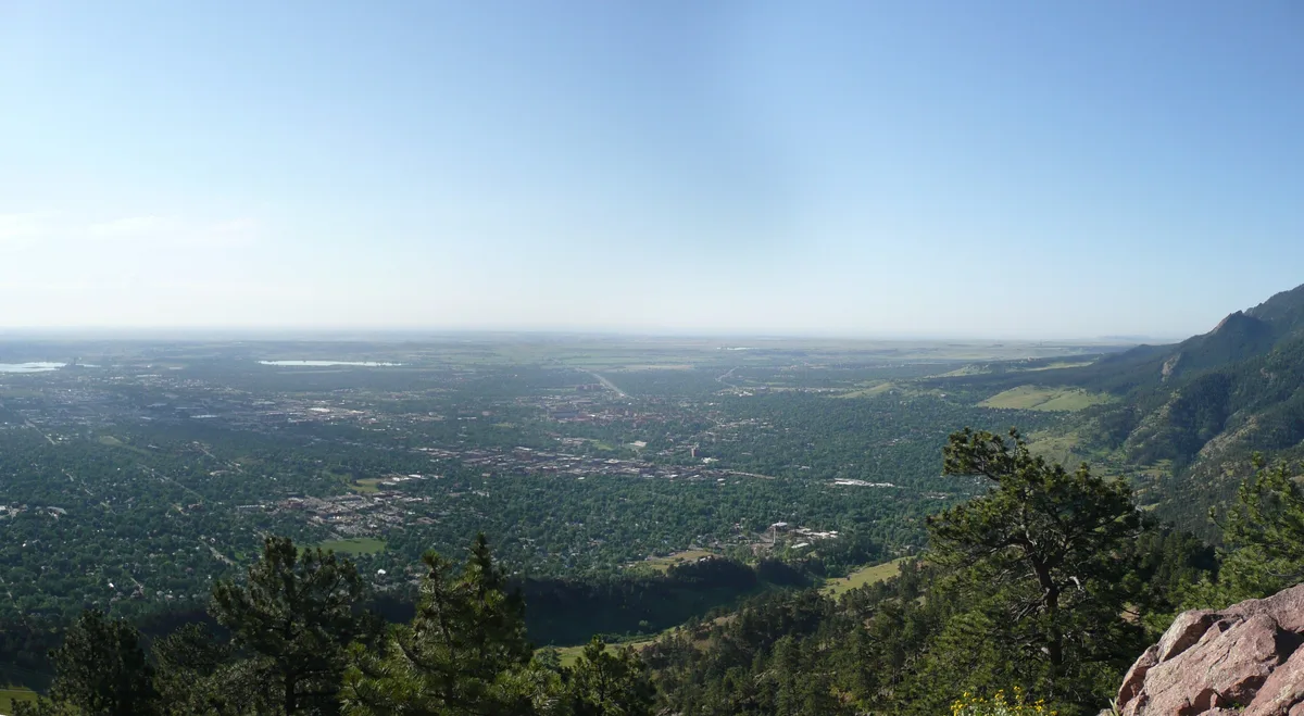 Wide panoramic view from Mount Sanitas over Boulder, with the city spreading across the valley toward the flat plains under a clear blue sky. Dark green foothills and pine trees frame the foreground, with sunlit rocky slopes and a boulder at the lower right.