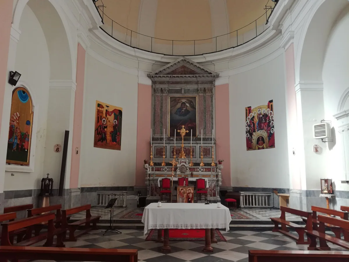 The image shows the interior of a small church sanctuary with a central altar, red chairs, and a checkered black-and-white tile floor. White and pale pink walls curve up to a domed ceiling, with colorful religious paintings and ornate marble detailing framing the altar.