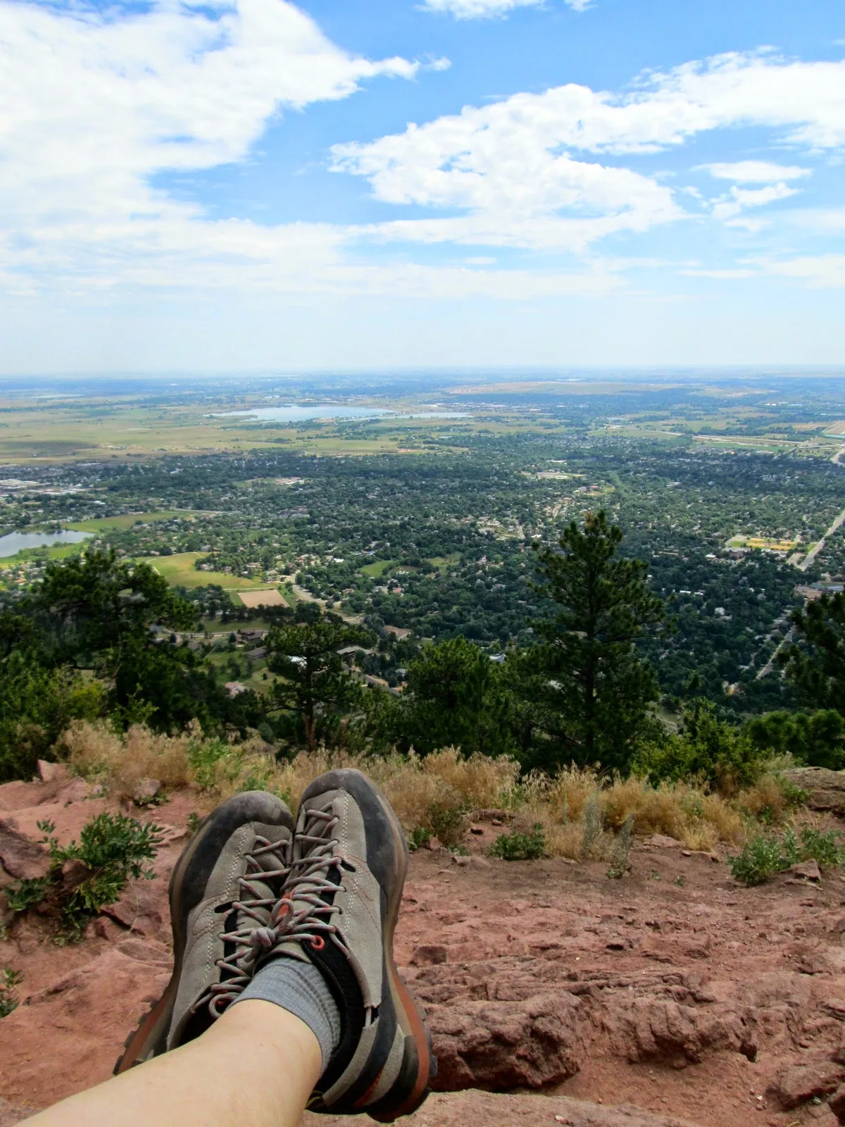 Boots rest on a rocky summit ledge above Boulder, with pine-covered foothills dropping into the city and the plains stretching to the horizon. A bright blue sky with scattered clouds fills the top half of the frame, giving the overlook a clear, airy feel.