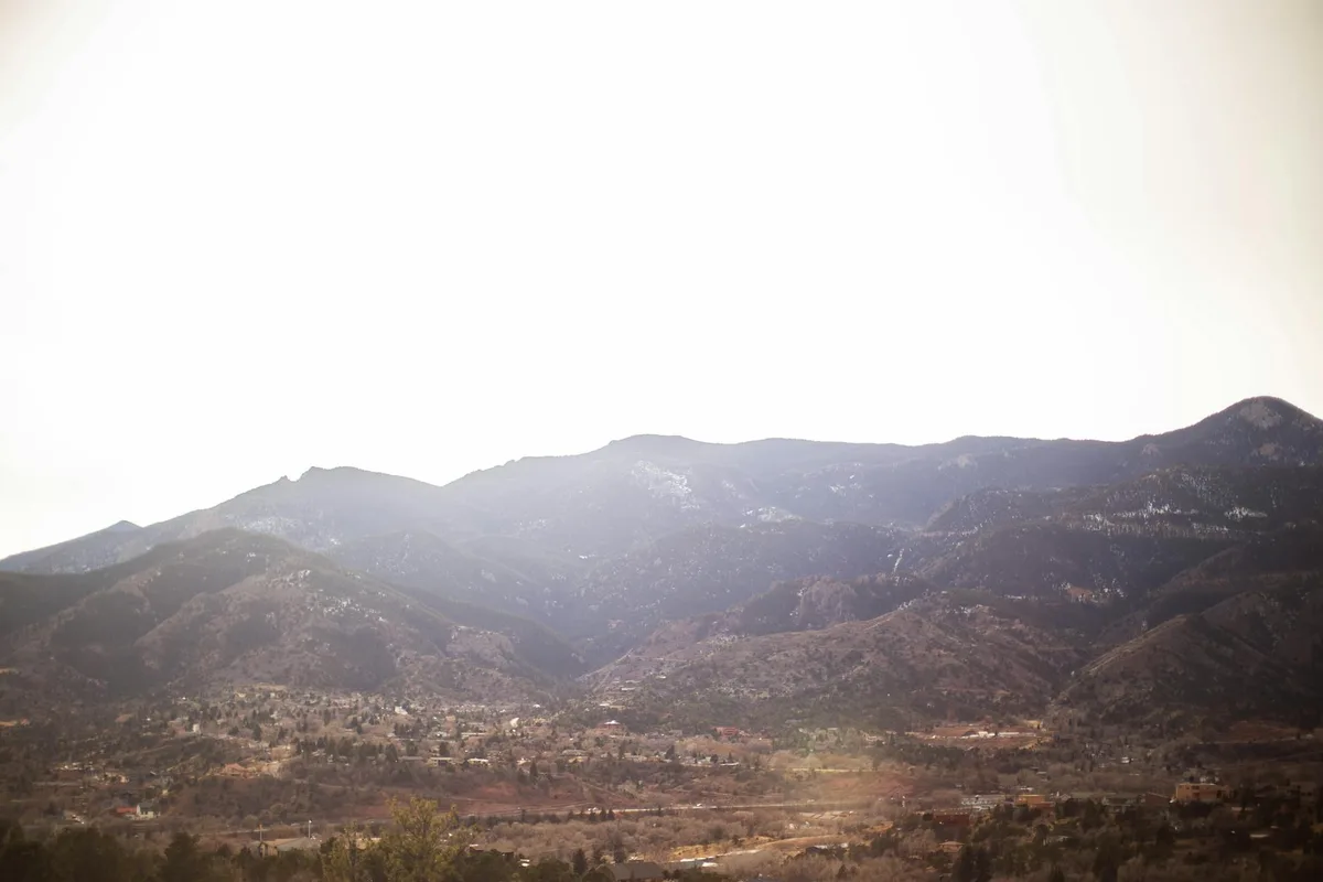 Bright golden-hour light washes over Boulder and the dark foothills, with a hazy mountain ridge stretching across the horizon. The city and valley sit below in soft detail, while the summit view feels wide and sunlit.