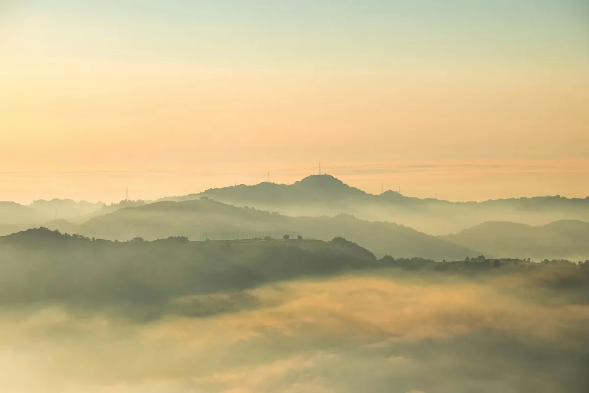 Golden-hour light washes over layered foothills and distant ridgelines, with soft fog pooling in the valleys below a pale orange sky. The scene feels calm and panoramic, with faint silhouettes of towers and hills fading into the mist.