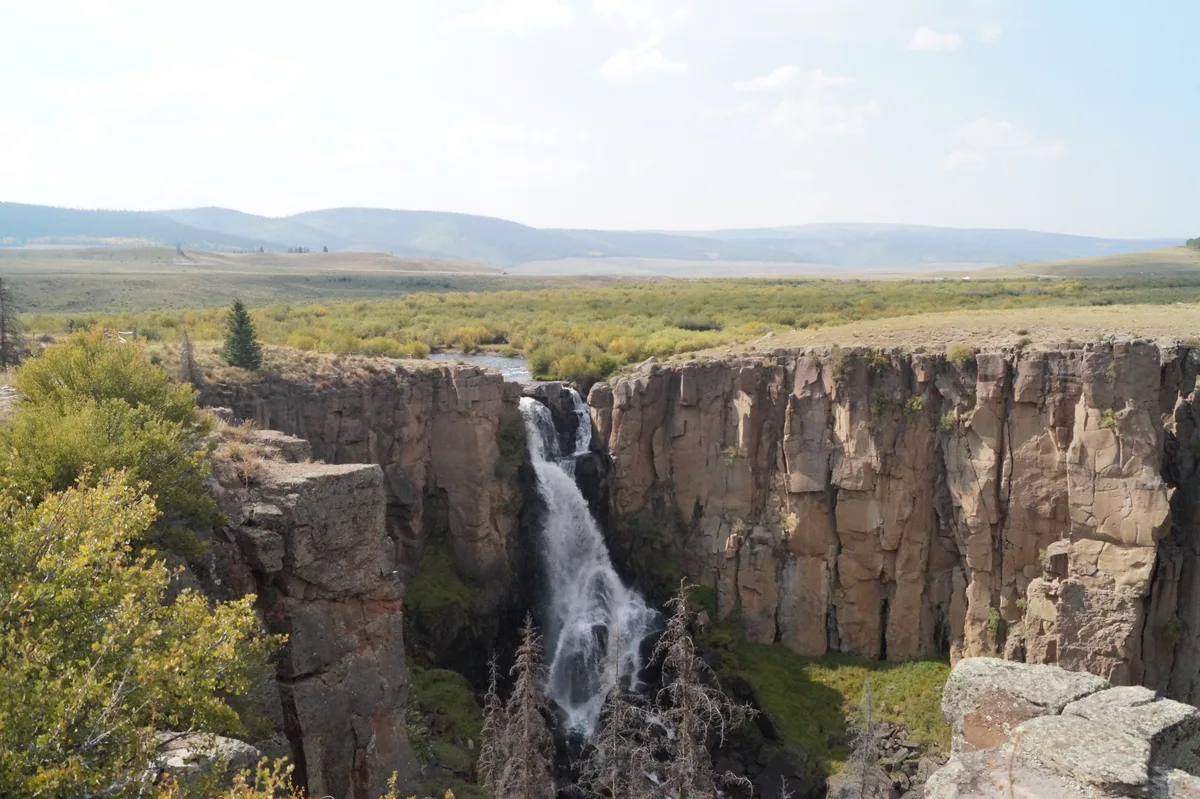 A tall waterfall spills over a sheer rhyolite cliff into a narrow rocky gorge, with misty white water contrasting against the dark chute below. Sunlit cliffs and sparse spruce and shrubs frame the falls, while open high-country hills stretch into the hazy distance.