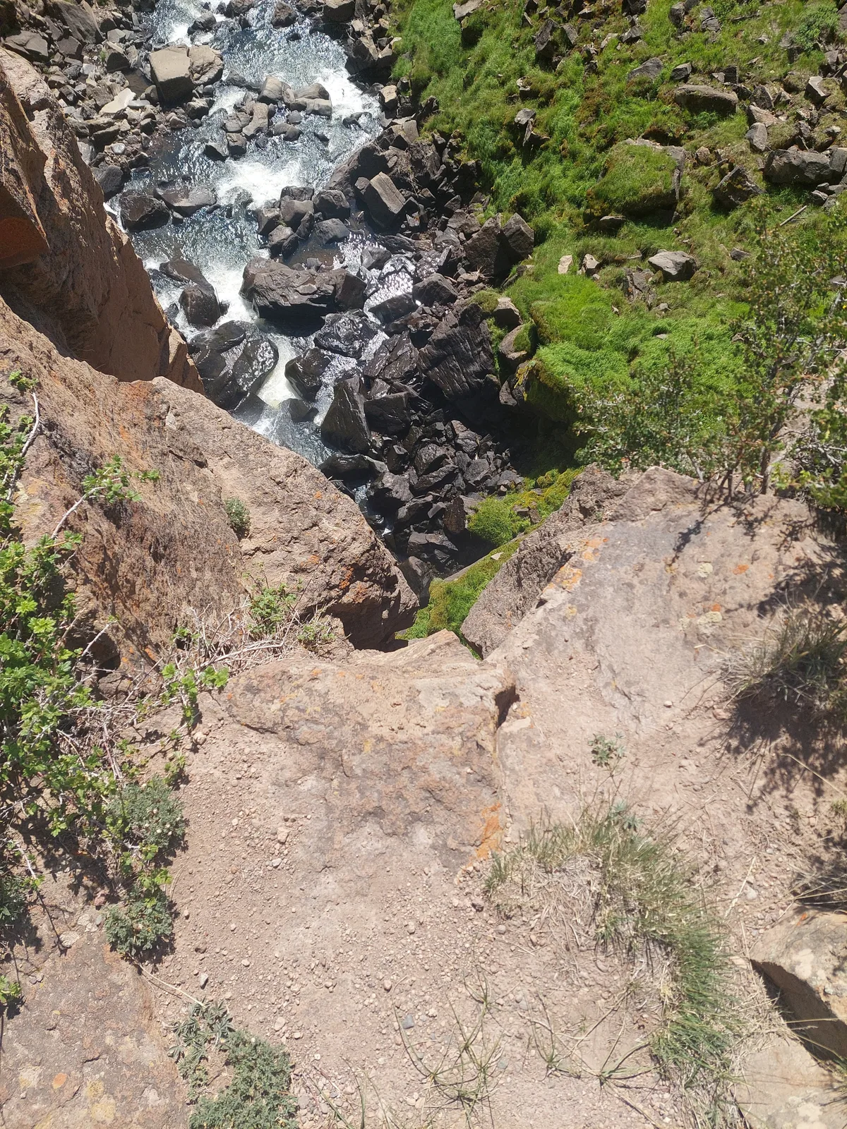 Looking straight down into a rocky gorge, white water rushes past dark boulders below tan rhyolite cliffs, with bright green moss and scrub clinging to the steep sides. Sunlit rocks and the narrow overlook ledge frame the falls on a clear morning.