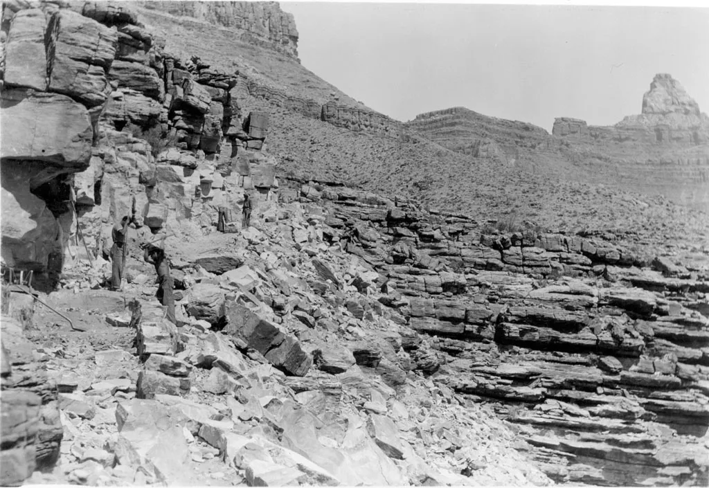 A black-and-white view of a rocky canyon wall and steep talus slope, with a few people standing along a rough trail carved into the cliffside. Layered stone ledges stretch across the gorge under a pale sky, giving the scene a stark, rugged feel.