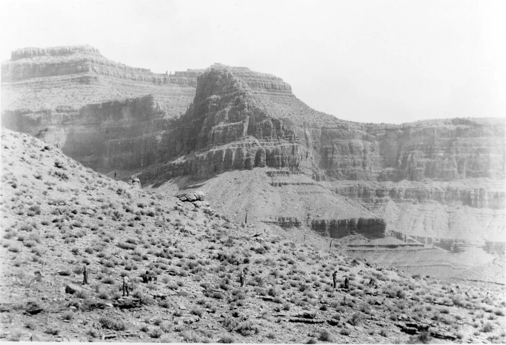 A black-and-white wide view of the Grand Canyon shows layered cliffs and a steep, scrub-covered slope in the foreground, with a few tiny figures scattered along the trail. Bright haze softens the distant rock walls and makes the central butte stand out against the pale sky.