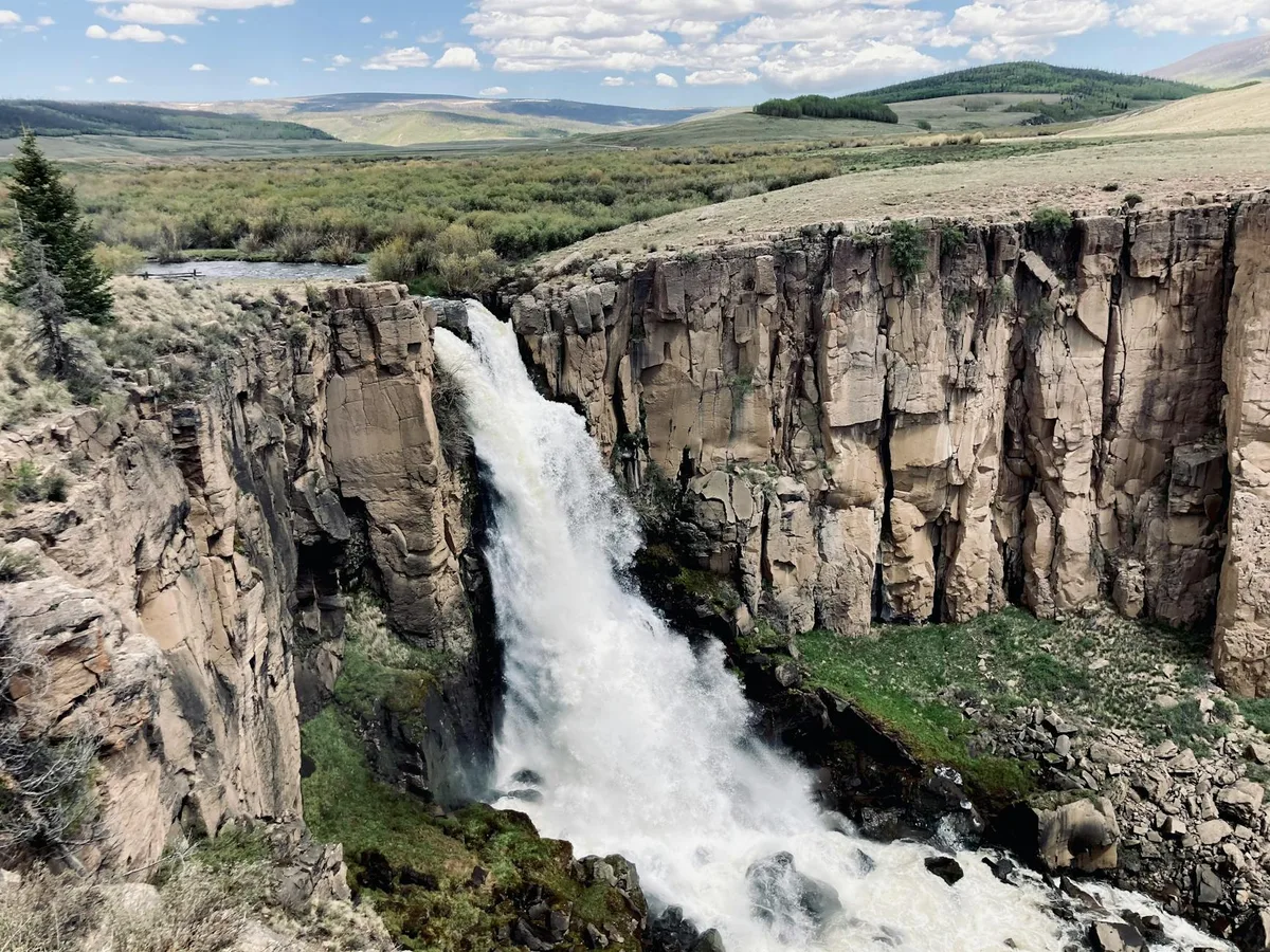 North Clear Creek Falls plunges in a thick white sheet over tall, tan rhyolite cliffs into a rocky gorge, with mossy green banks below. Rolling high-country meadows and distant hills stretch beyond the falls under a bright morning sky with scattered clouds.
