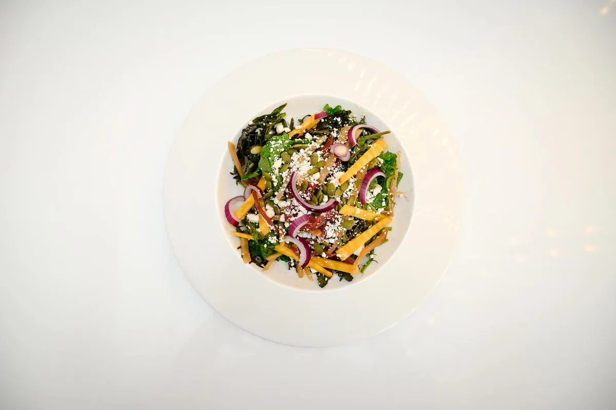 A top-down view of a colorful salad on a white plate against a bright white background, with red onion rings, shredded cheese, seeds, and leafy greens scattered across the center. The clean, minimal composition leaves lots of negative space around the dish.
