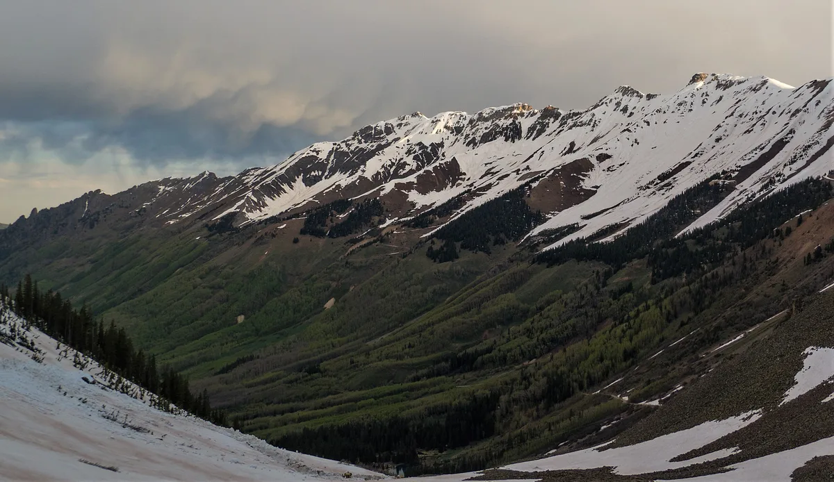 Snow-striped alpine peaks rise above dark green forested slopes, with a broad valley dropping away below under a muted morning sky. The foreground shows a steep, rocky pass edge with lingering snow patches, emphasizing the high, remote terrain.