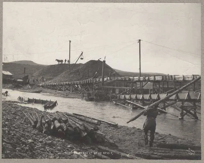 Historic mining works line a narrow stream, with wooden sluices, gravel piles, and rough log supports stretching across the water under a pale sky. A few small figures and a horse stand on the ridge, emphasizing the remote, early-1900s industrial scene.