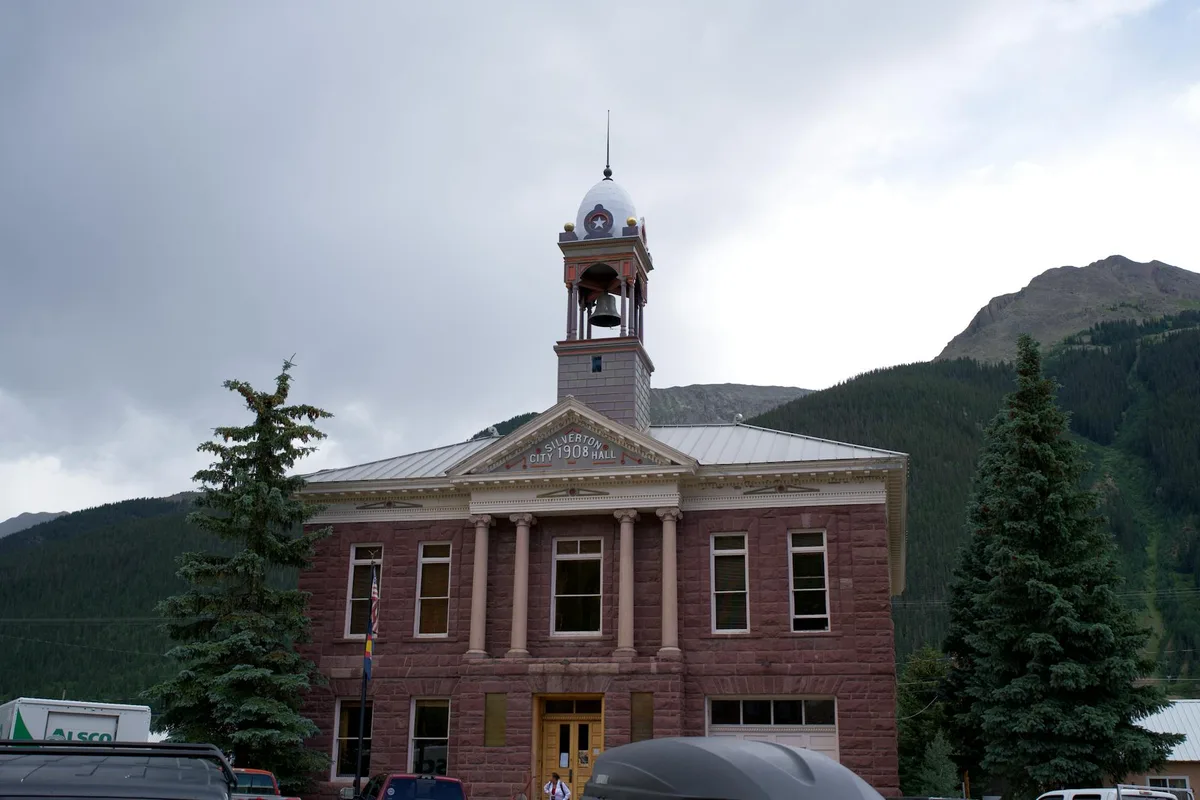 Silverton City Hall stands centered in the frame, its pink stone facade and clocktower bell set against dark green mountain slopes and a pale morning sky. Tall evergreens flank the historic building, with parked vehicles at street level adding a small-town feel.