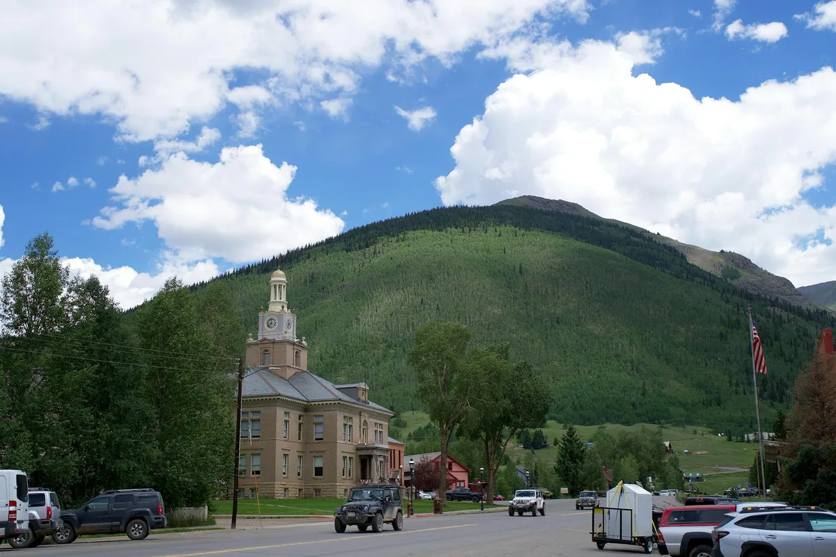 A small mountain town street sits below a steep green hillside under a bright blue sky with puffy white clouds. Historic buildings, parked vehicles, and a clock-tower courthouse line the road, with tall trees framing the scene.