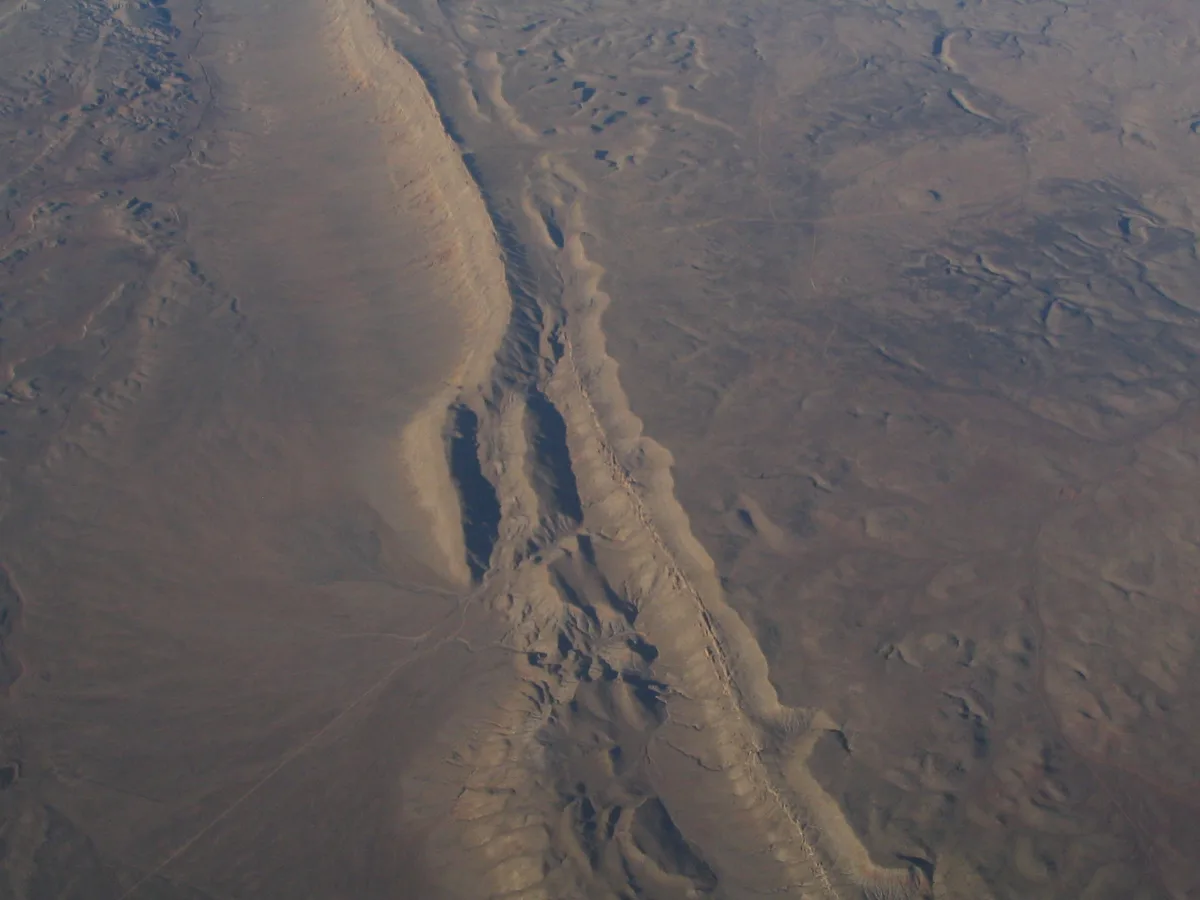 A wide, hazy aerial view shows a long rocky ridge and eroded desert slopes, with warm golden light catching the crest and casting deep shadows into the gullies. The terrain is mostly muted brown and gray, textured with ridgelines, washes, and sparse scrub.