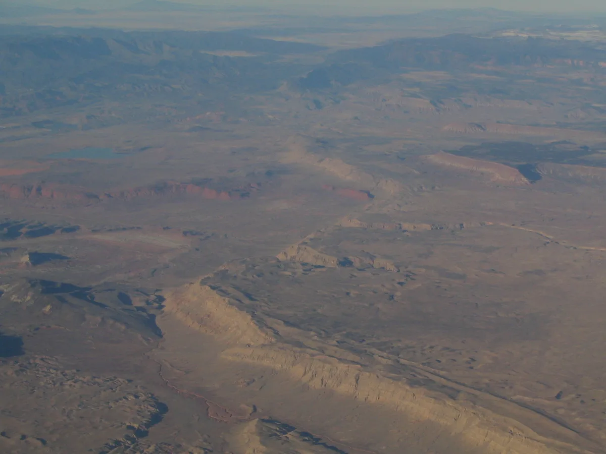 Sunlit tan cliffs and ridgelines stretch across a broad desert valley, with long blue mountain shadows fading into the hazy distance. The golden-hour light picks out layered rock formations and dry washes, creating a soft, panoramic view.